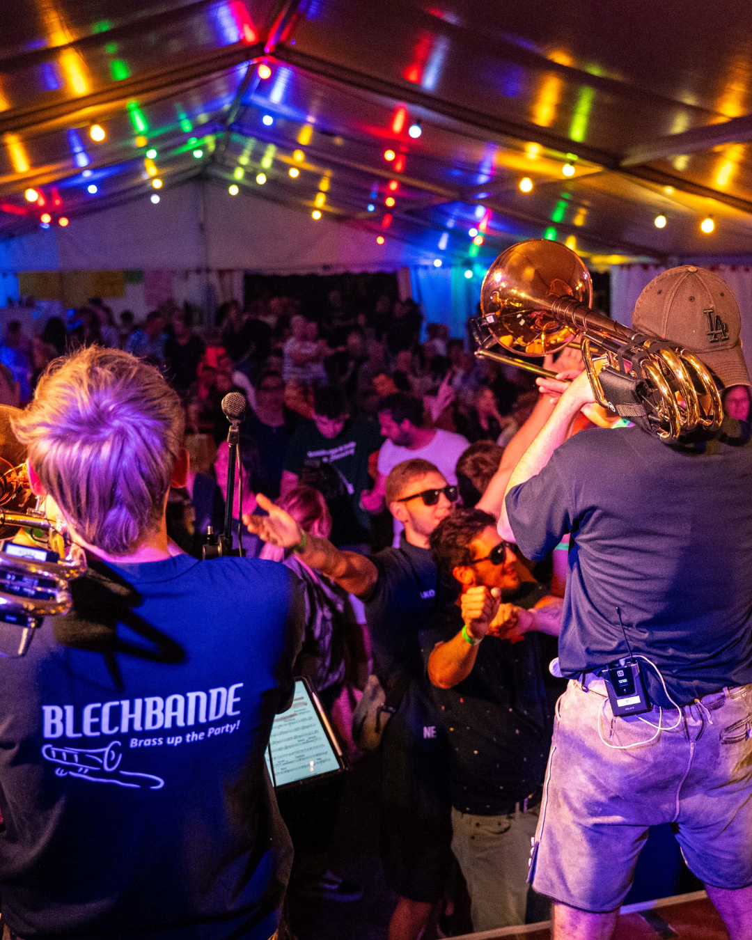 Menschen tanzen und feiern unter bunten Lichtern in einer festlichen Zeltveranstaltung, während Musiker auf einer Bühne spielen.
