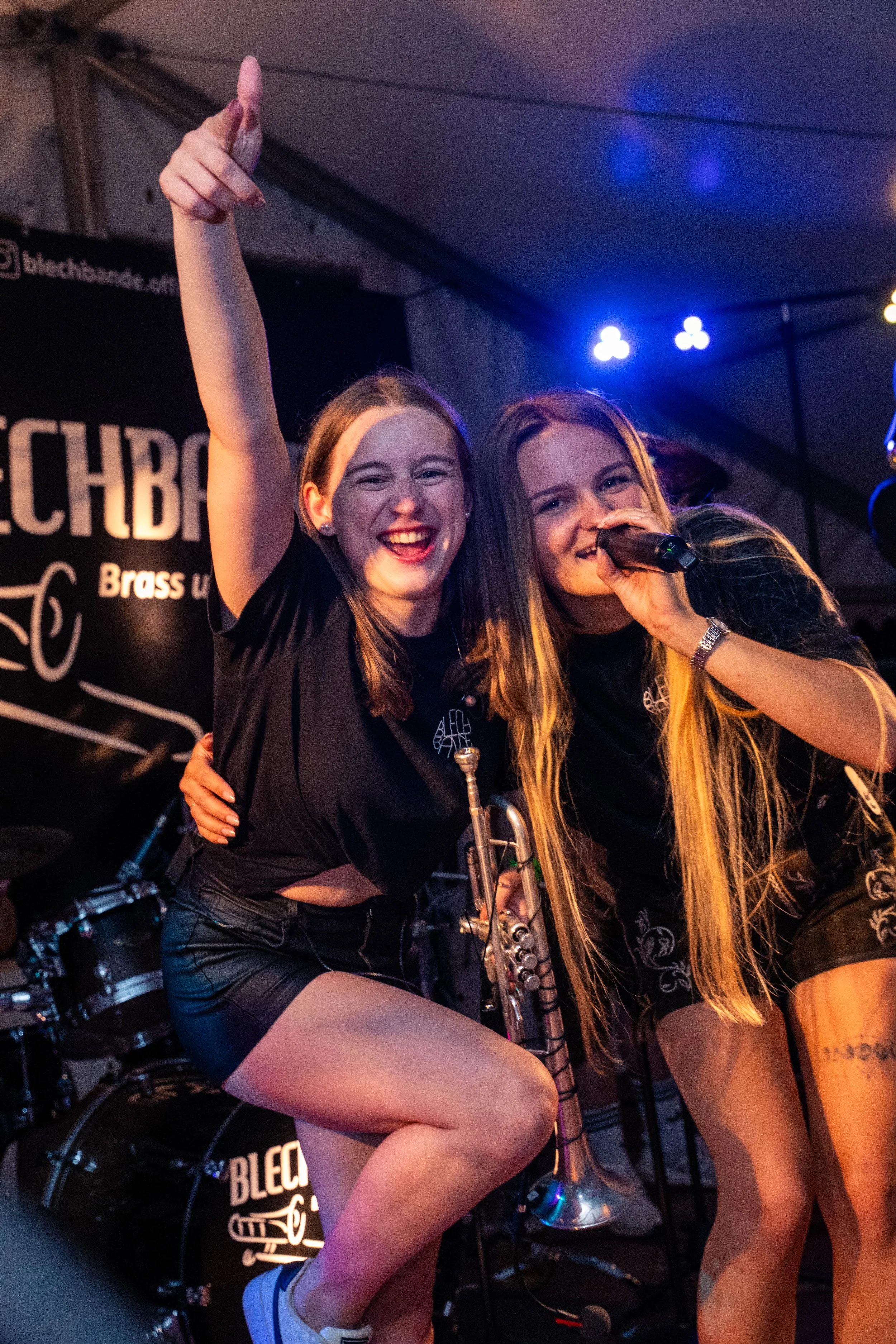 Zwei lachende junge Frauen bei einem Musikauftritt, eine singt in ein Mikrofon, die andere hebt die Hand in die Luft, im Hintergrund eine Bühne mit Schlagzeug und einer schwarzen Band-Branding