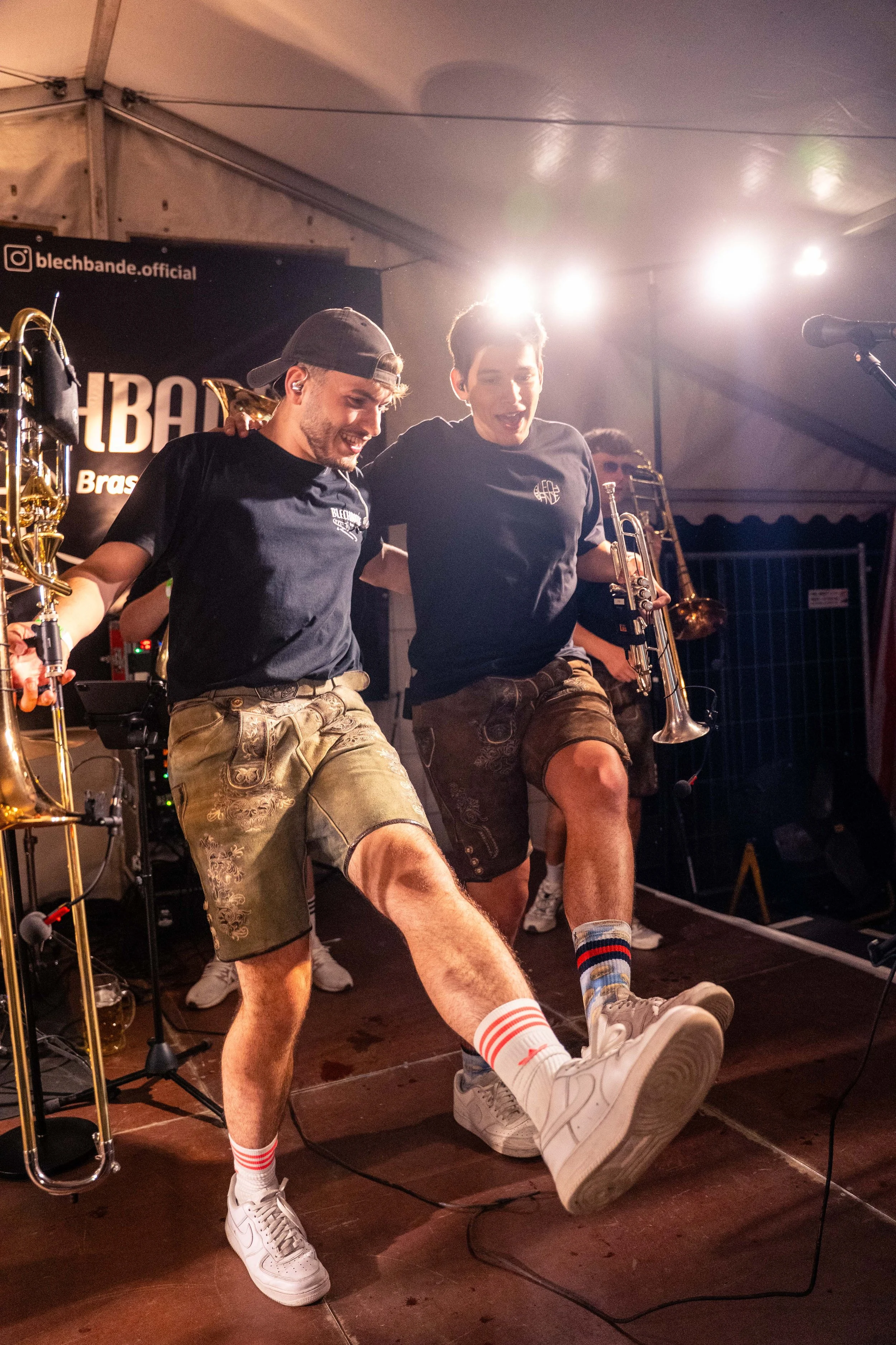 Zwei Männer in traditionellen bayerischen Lederhosen tanzen auf einer Bühne bei einem Musikfestival, während sie Musikanten begleiten.