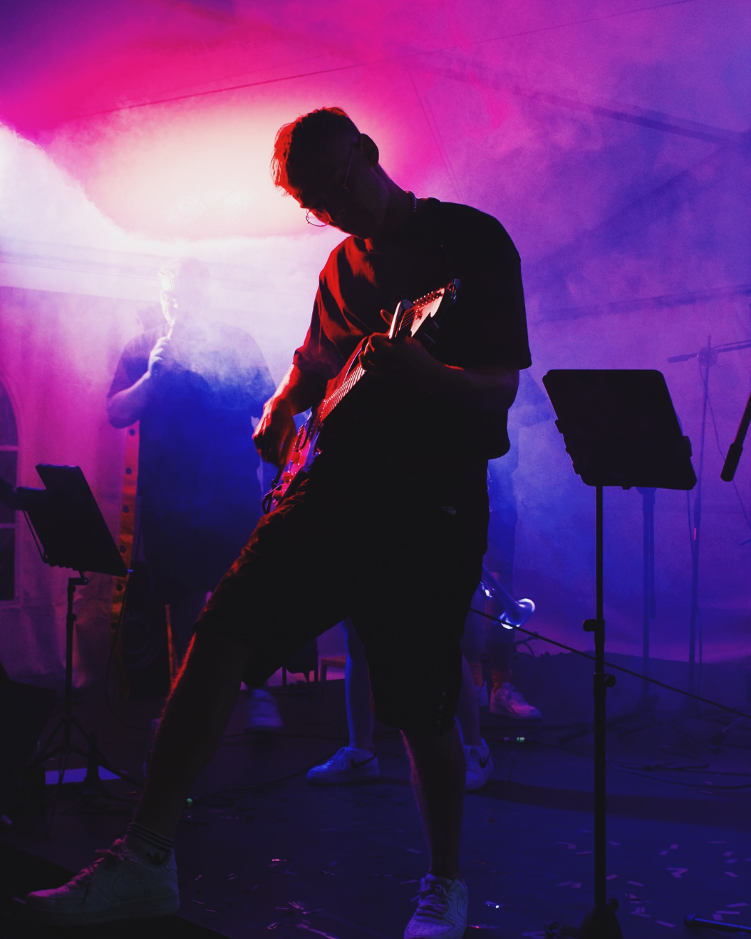 Silhouette eines jungen Mannes, der Gitarre spielt, umgeben von buntem Bühnenlicht in Pink und Blau, auf einer Bühne während eines Live-Auftritts.