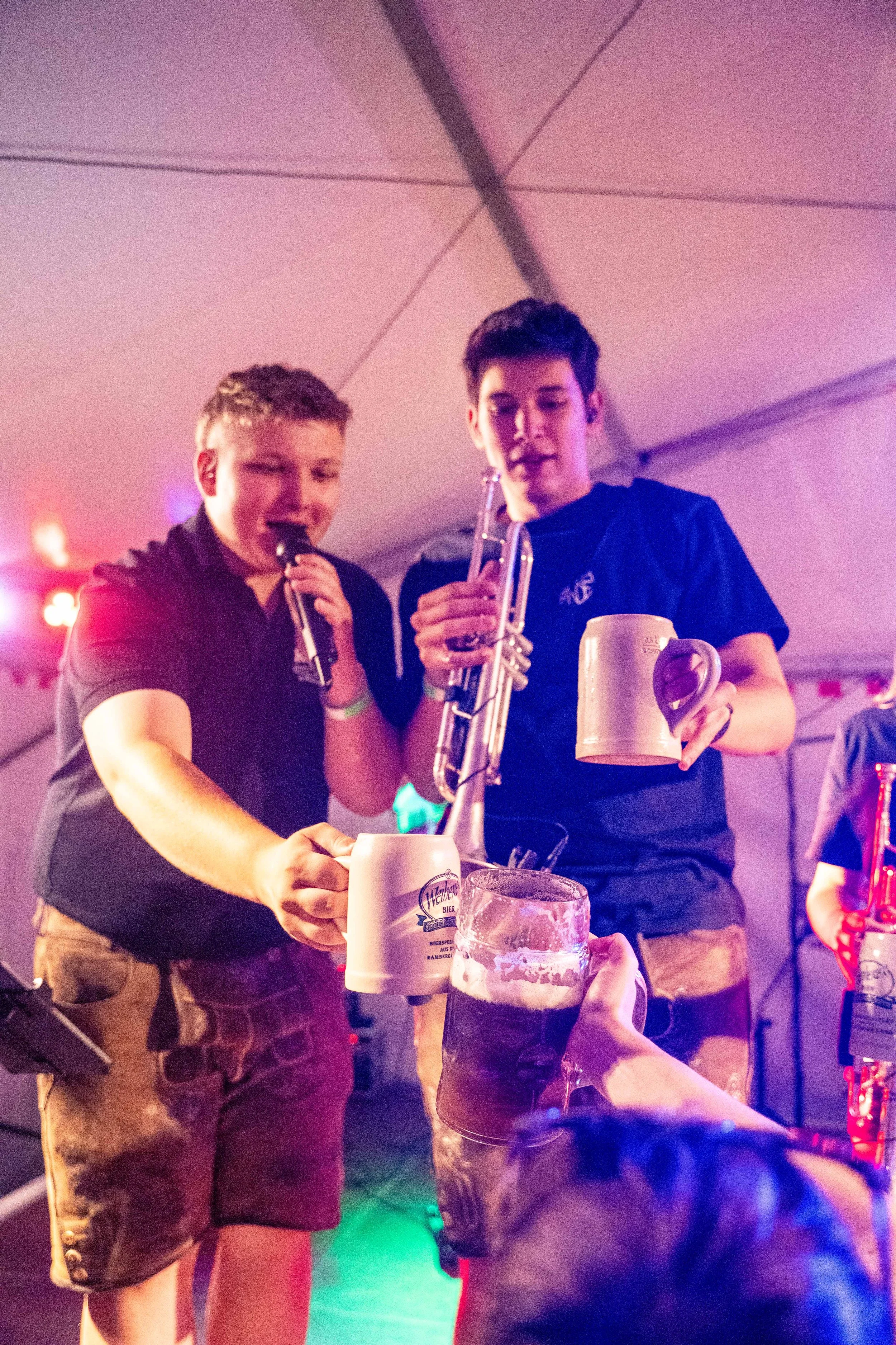 Zwei junge Männer in einem Zelt bei einem Fest, die Bierkrüge hochhalten und anstoßen.