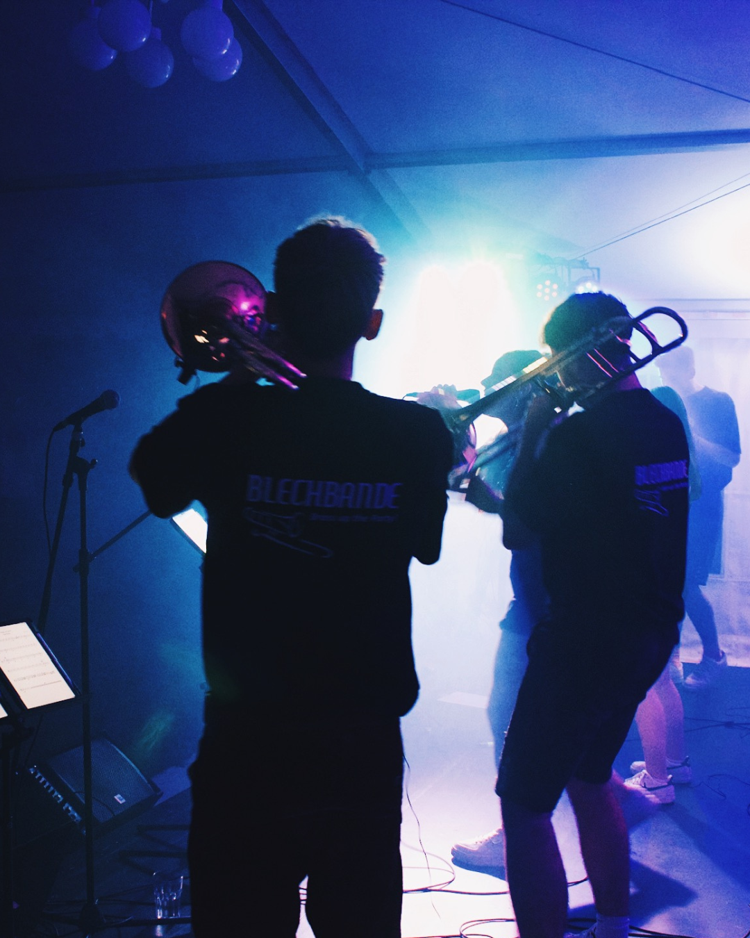 Musiker spielen auf einer Bühne mit bunten Lichtern, einer hält eine Trompete, im Hintergrund andere Konzertbesucher.