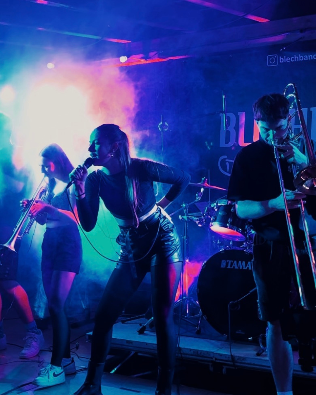 Jugendliche Musikerinnen und Musiker spielen auf einer Bühne, umgeben von bunten Lichteffekten, mit einem Schlagzeug im Hintergrund.