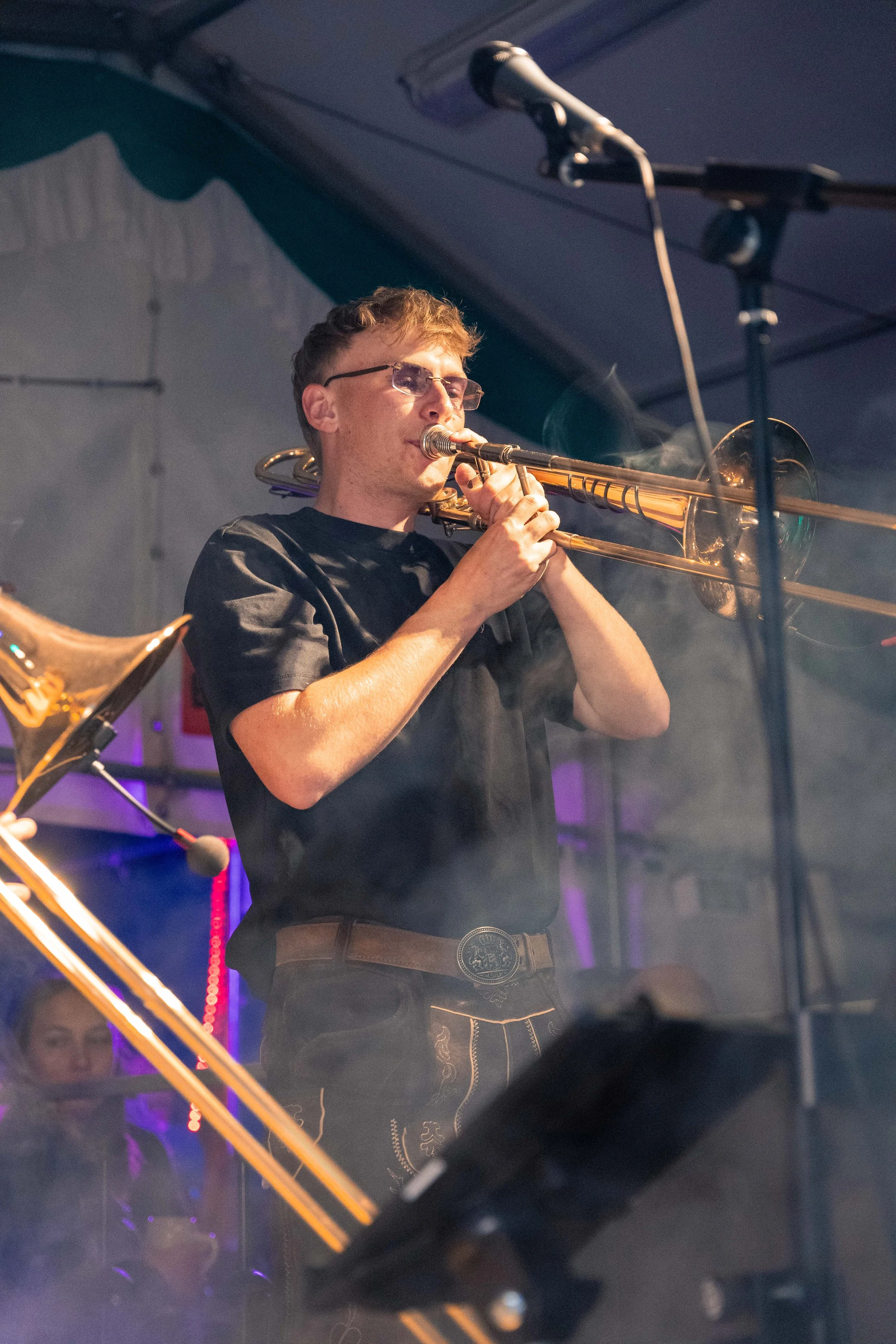 Ein junger Mann spielt  Posaune auf einer Bühne, trägt Sonnenbrille und dunkle Kleidung, mit Mikrofon im Hintergrund.