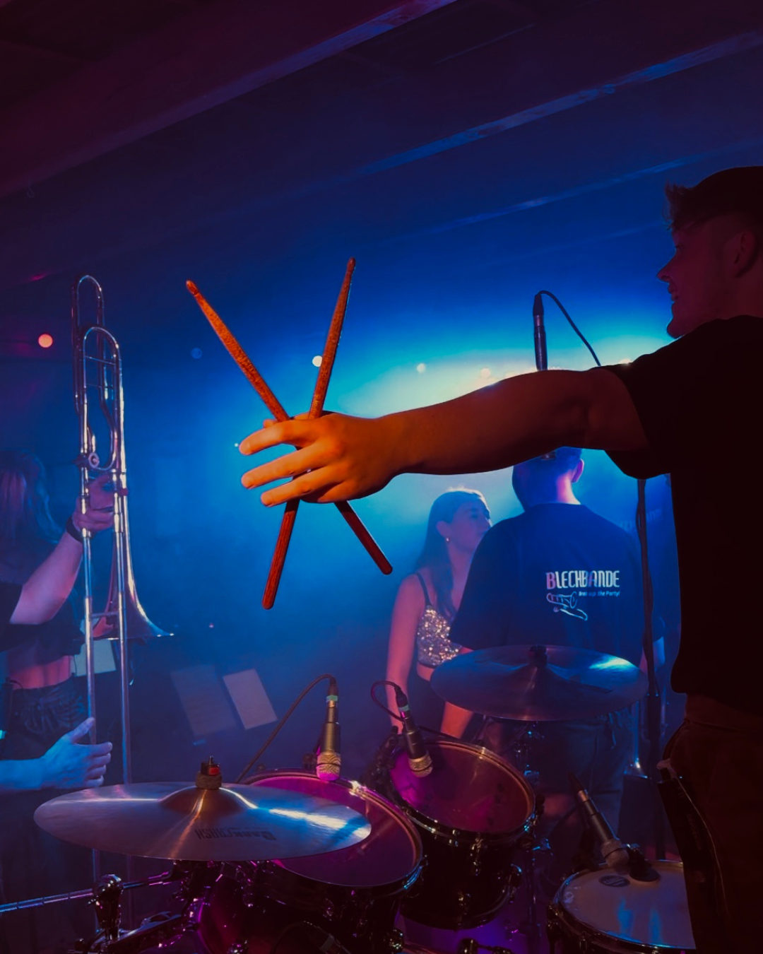 Musiker auf der Bühne während eines Live-Auftritts, mit Farblich beleuchteter Atmosphäre, einem Schlagzeug und einer Hand, die Schlagzeugsticks hält, im Vordergrund.