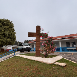 Cena ao ar livre de uma árvore de grande porte com folhas verdes, uma cruz de Jesus Cristo e uma casa de telhado de cerâmica ao fundo, céu azul com algumas nuvens, e gramado verde no primeiro plano.