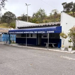 Prédio de unidade de saúde municipal de Cotia com fachada azul, toldo azul com escritural 'U NERÁRIO MUNICIPAL DE COTIA 24hrs' e interiores visíveis pela porta aberta, com plantas decorativas na entrada.