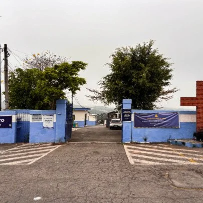 Entrada de uma escola com muralha azul e portões abertos, árvores ao fundo e carro estacionado na rua.