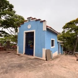 Imagem de uma pequena capela com parede cinza, porta azul com grades, e uma cruz branca acima. No topo, há uma placa com o texto 'COTIA'.
