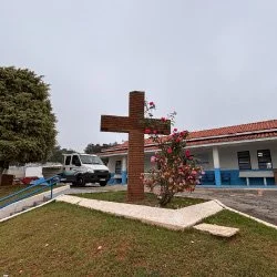 Cena ao ar livre de uma árvore de grande porte com folhas verdes, uma cruz de Jesus Cristo e uma casa de telhado de cerâmica ao fundo, céu azul com algumas nuvens, e gramado verde no primeiro plano.
