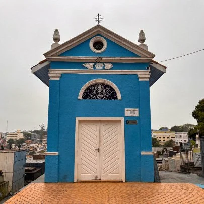 Capela azul com porta branca, detalhes arquitetônicos clássicos e cruz no topo, localizada em um cemitério.