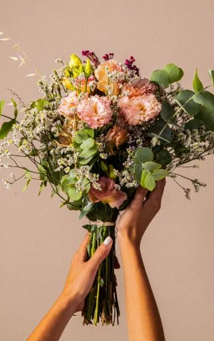 Mãos segurando um buquê de flores com rosa, branco, amarelo e roxo, e folhas verdes contra um fundo bege.