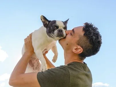 Homem segurando um cachorro Boston Terrier, que está sendo beijado na face. Fundo de céu azul claro.
