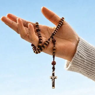 Mãos segurando um terço com uma cruz, contra um céu azul claro.