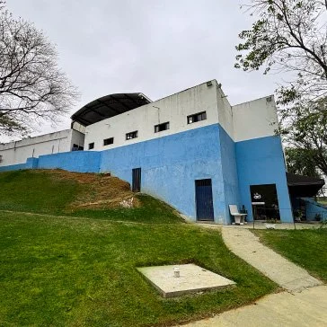Prédio azul claro com partes brancas, situado em uma colina com gramado e árvores ao redor, com acesso por uma rampa de concreto.