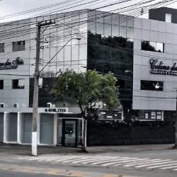 Recepção de um estabelecimento chamado Colina dos Ipês, que é um funerária, planearia e velório. Na recepção, duas mulheres estão sentadas atrás do balcão de madeira, uma delas ao lado de uma pilha de embrulhos de presente. No fundo, uma tela de TV, uma planta e alguns livros ou folhetos na mesa ao lado.
