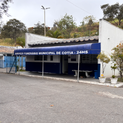 Prédio de unidade de saúde municipal de Cotia com fachada azul, toldo azul com escritural 'U NERÁRIO MUNICIPAL DE COTIA 24hrs' e interiores visíveis pela porta aberta, com plantas decorativas na entrada.