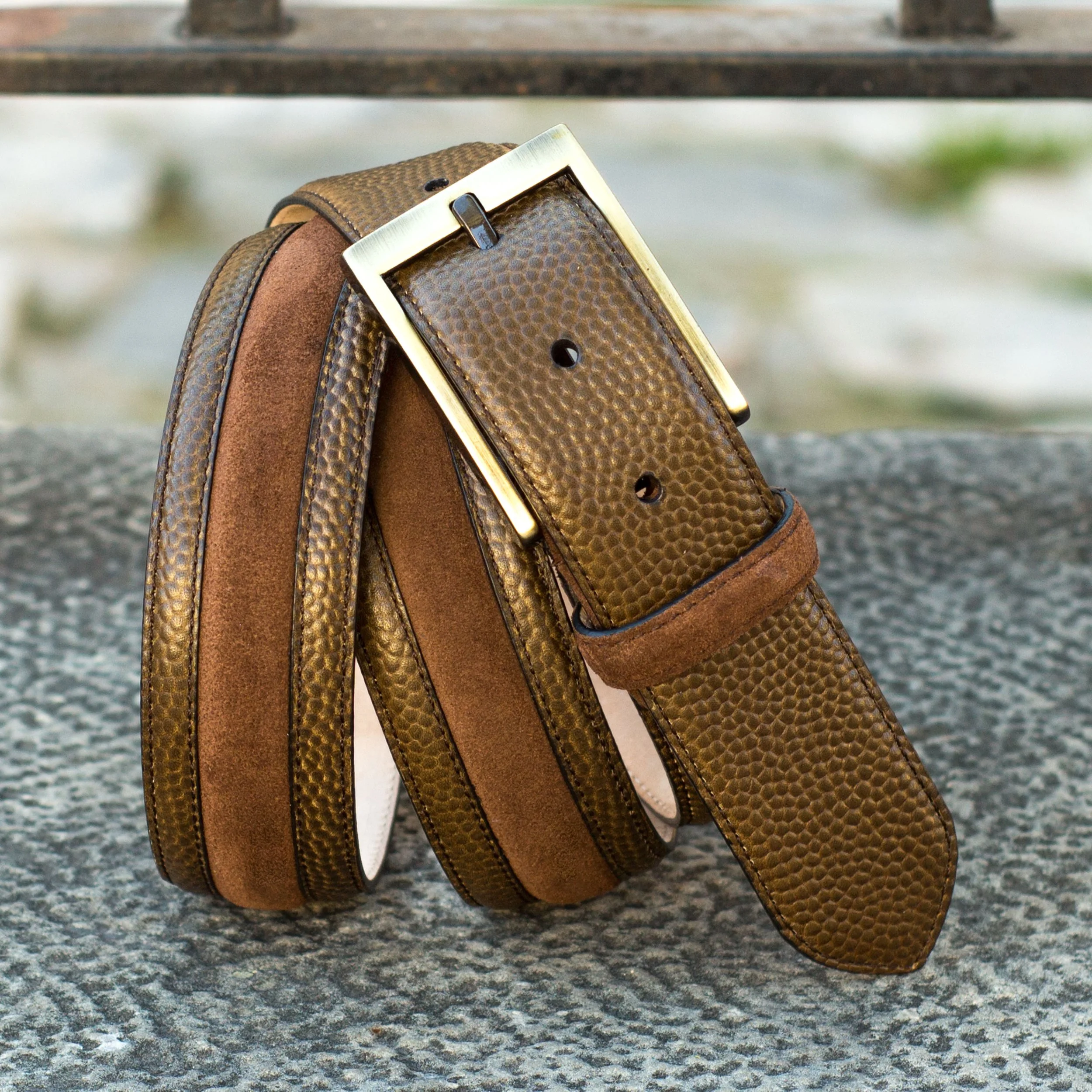 Brown leather and suede belt with gold buckle placed on a textured gray surface with a blurred outdoor background.