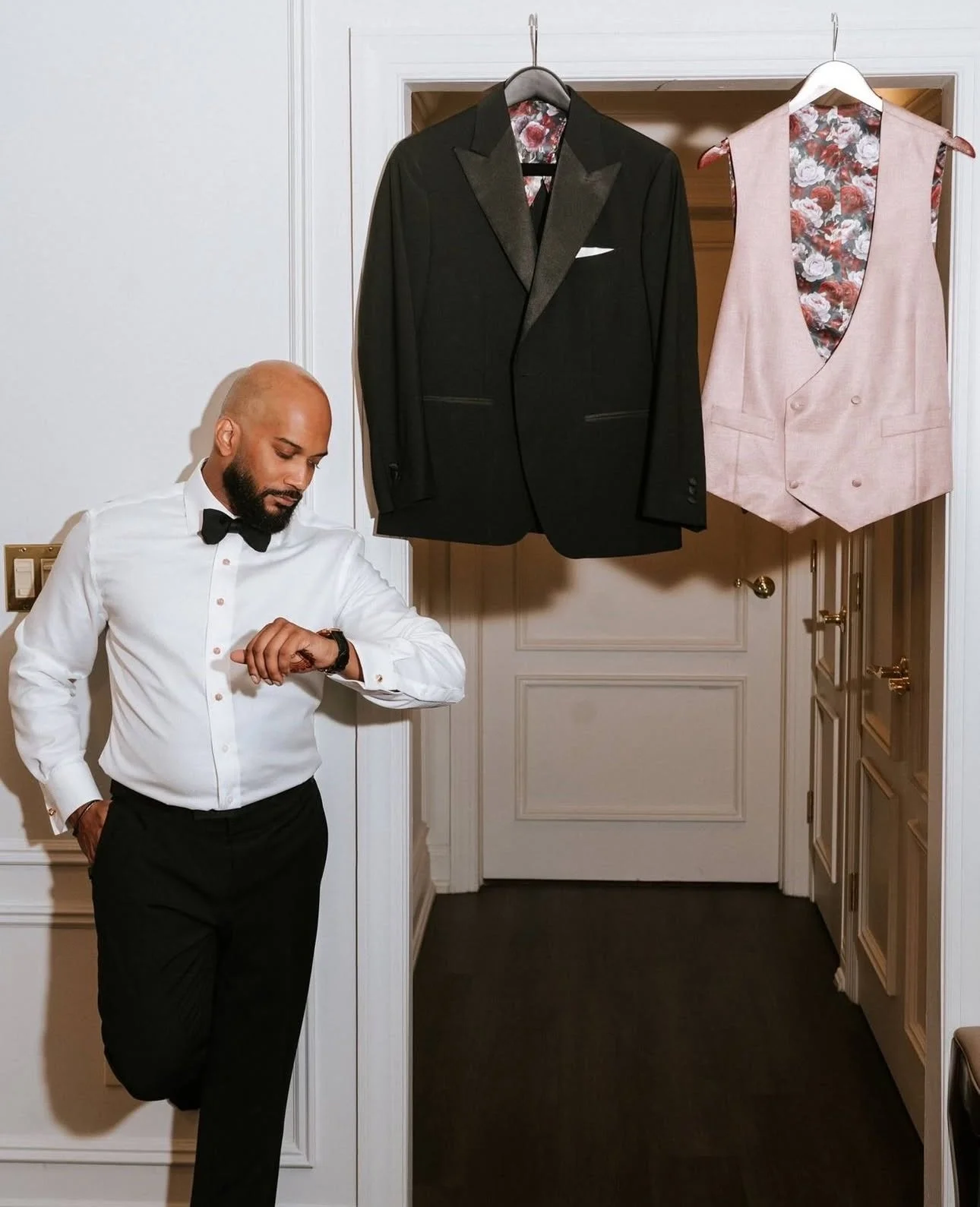A man in a tuxedo looking at his watch in a hallway, with two suits hanging on the door behind him, one black tuxedo and one pink vest with floral lining.