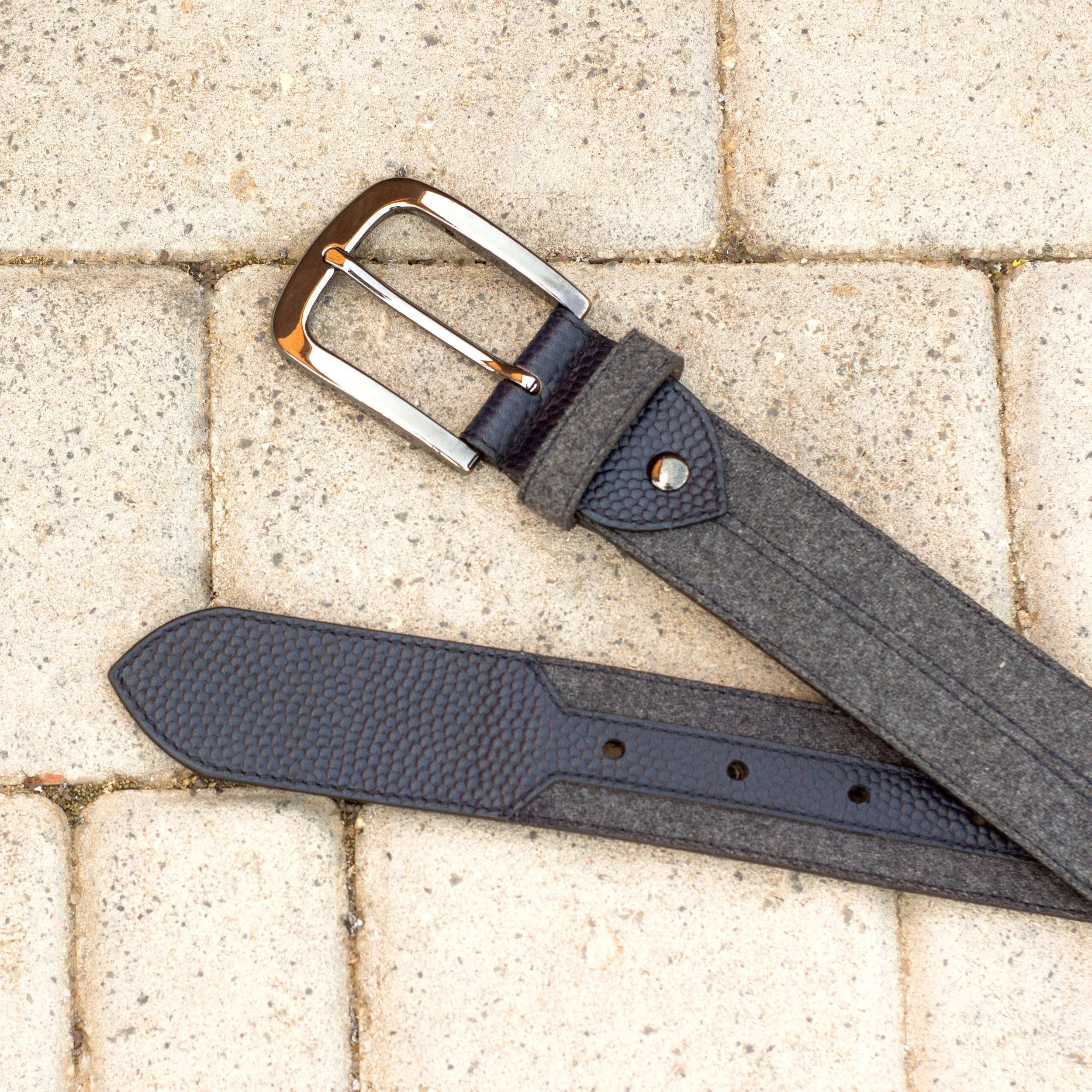A pair of black belts with textured leather at the tips, one with a shiny metal buckle, lying on light-colored paving stones.
