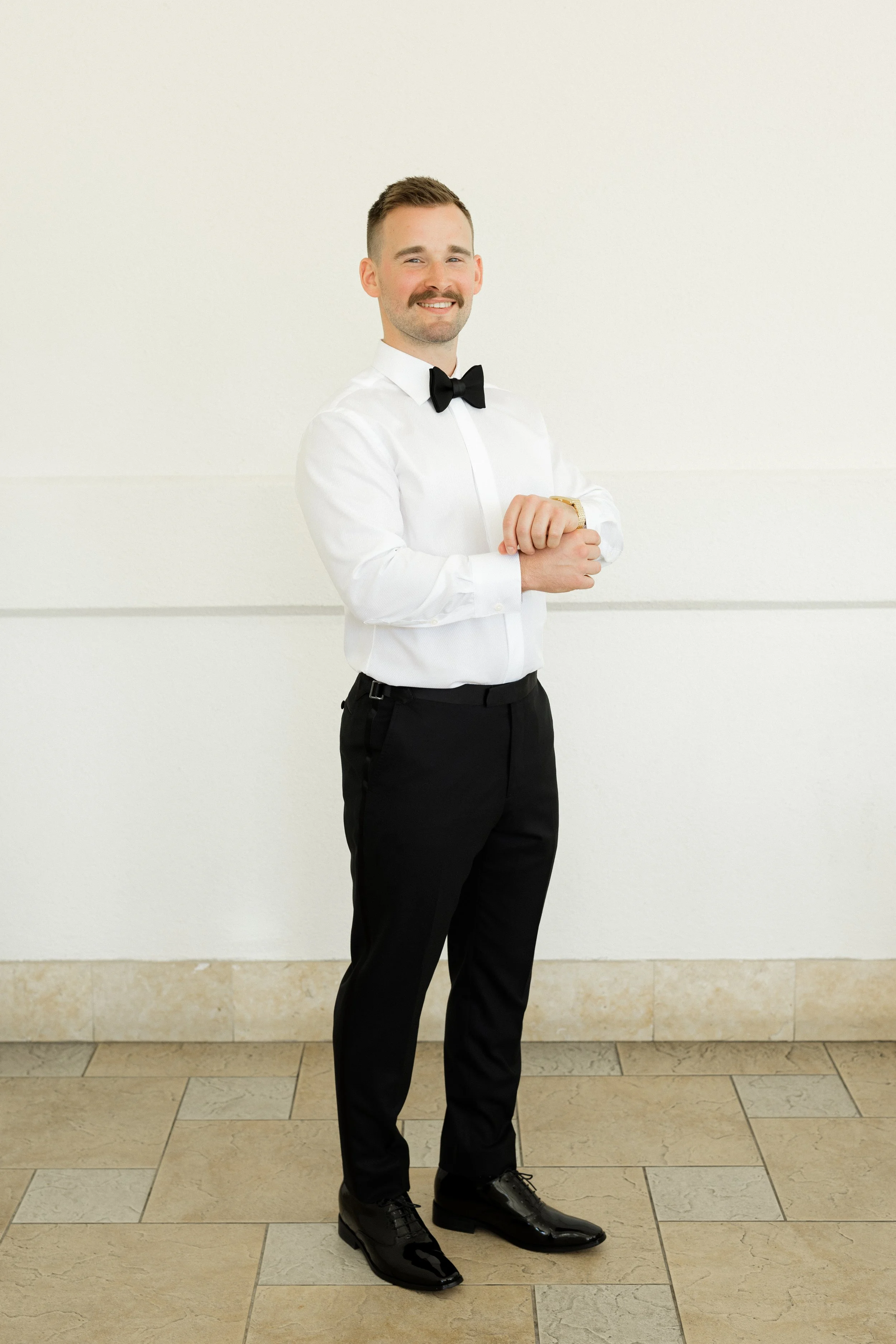 A man in a white dress shirt, black dress pants, black dress shoes, and a black bow tie standing against a white wall, smiling and looking at the camera.