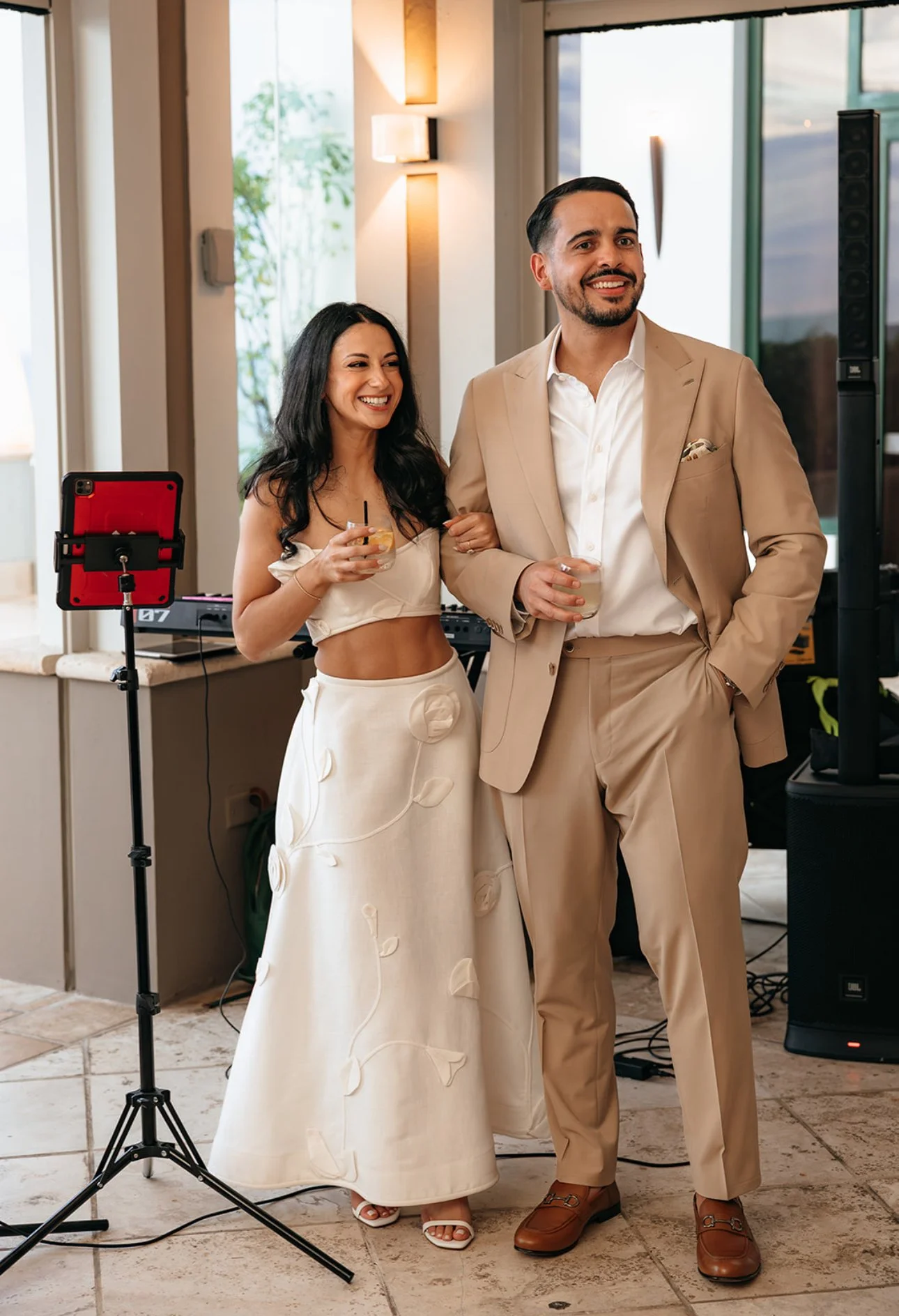 A man and woman in formal attire smiling at a social gathering, holding drinks, with a DJ setup in the background.