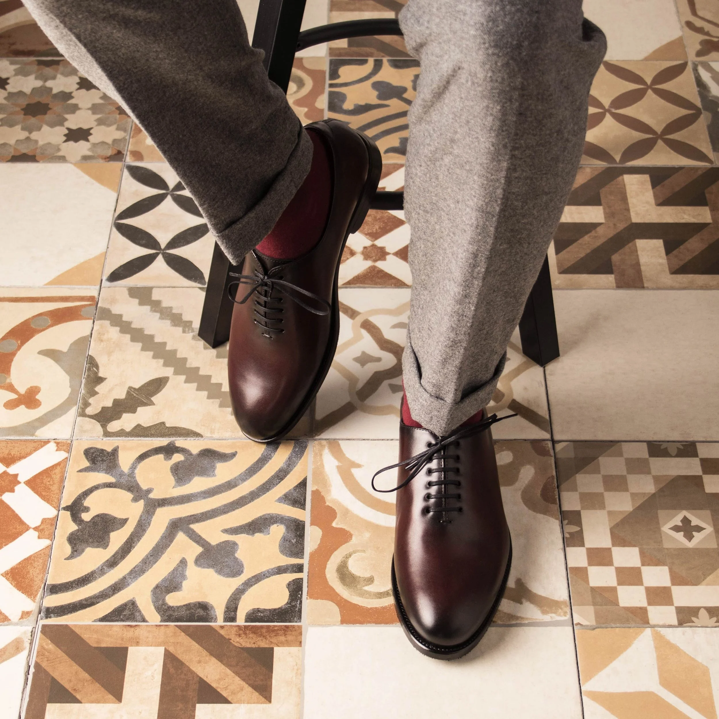 Person wearing dark polished dress shoes, gray pants, and red socks, sitting on a black chair on a tiled floor with various patterned tiles.