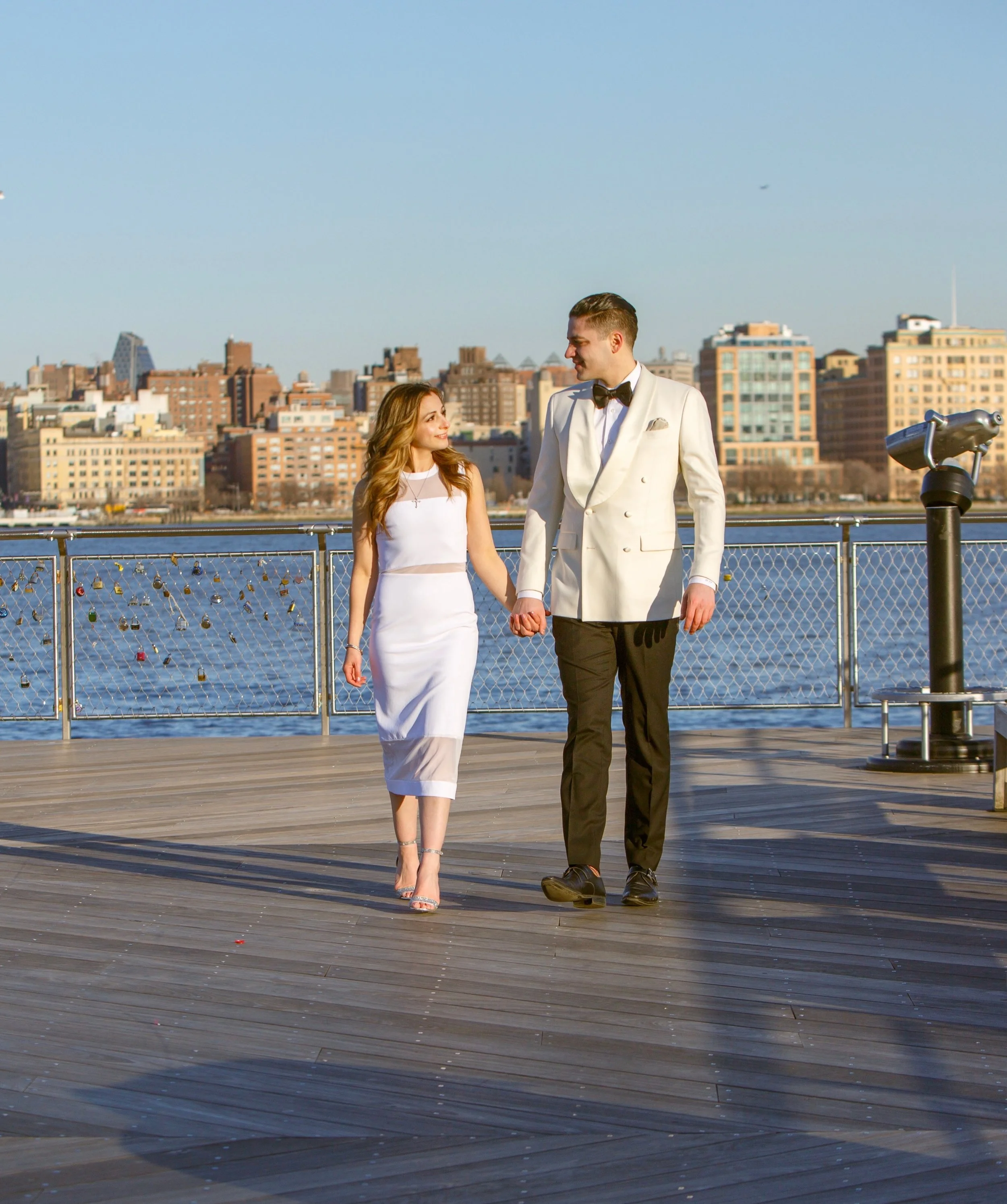 A young couple dressed in wedding attire holding hands and walking on a boardwalk near a body of water with a city skyline in the background.