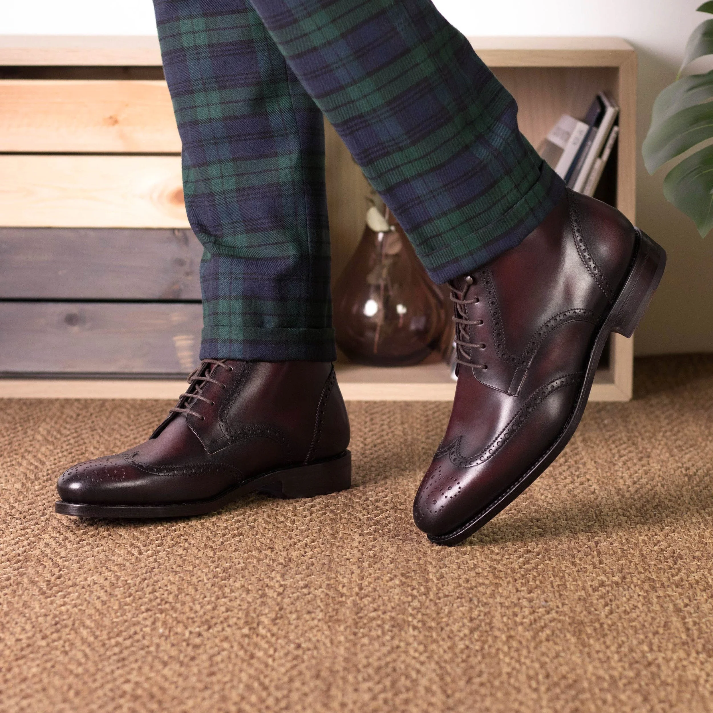 Person wearing dark brown leather brogue dress shoes, green and navy tartan plaid pants, standing on beige carpeted floor in front of a wooden shelf with books and decor.