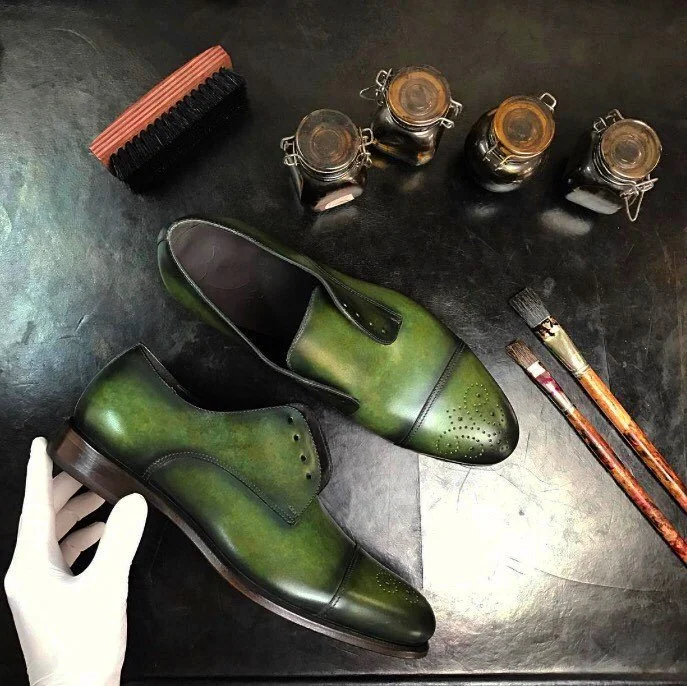 A pair of green leather dress shoes, three brushes, four small jars with lids, a shoe polishing sponge, and two paintbrushes on a black surface, with a gloved hand holding one shoe.