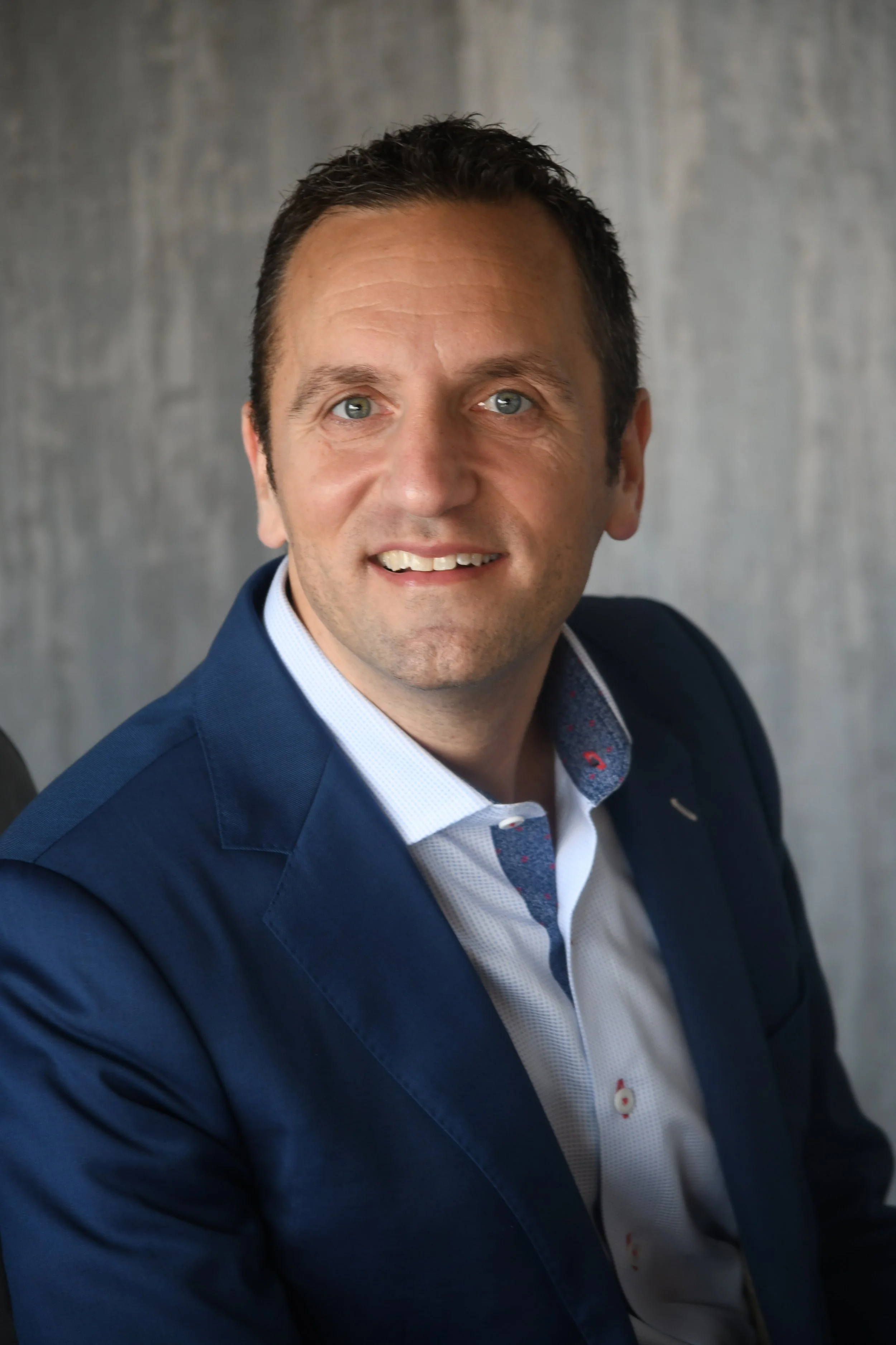 A man with dark hair and blue eyes wearing a blue blazer and a white shirt, smiling, against a gray background.