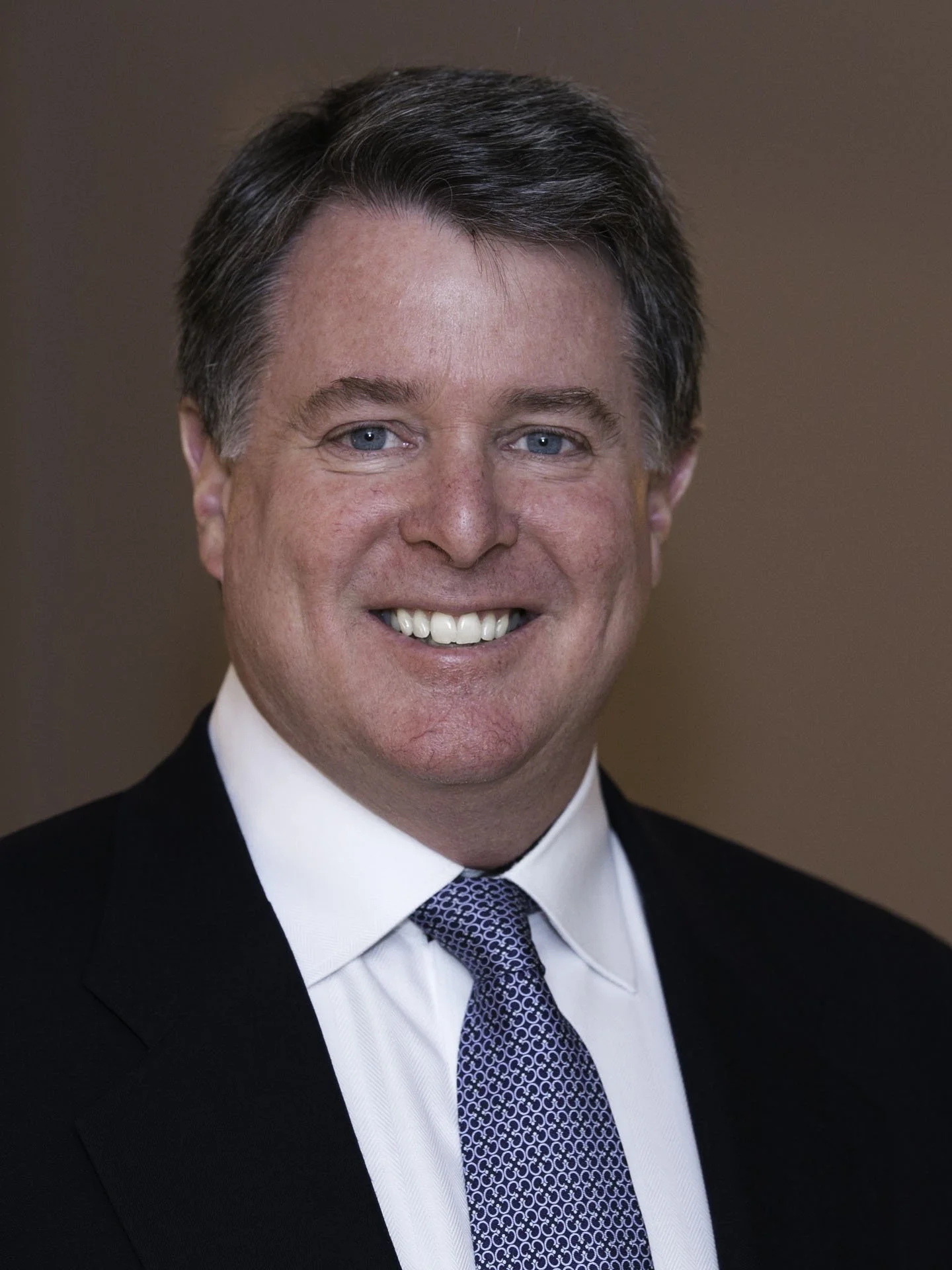 A smiling man with dark hair, blue eyes, wearing a black suit, white shirt, and patterned blue tie.
