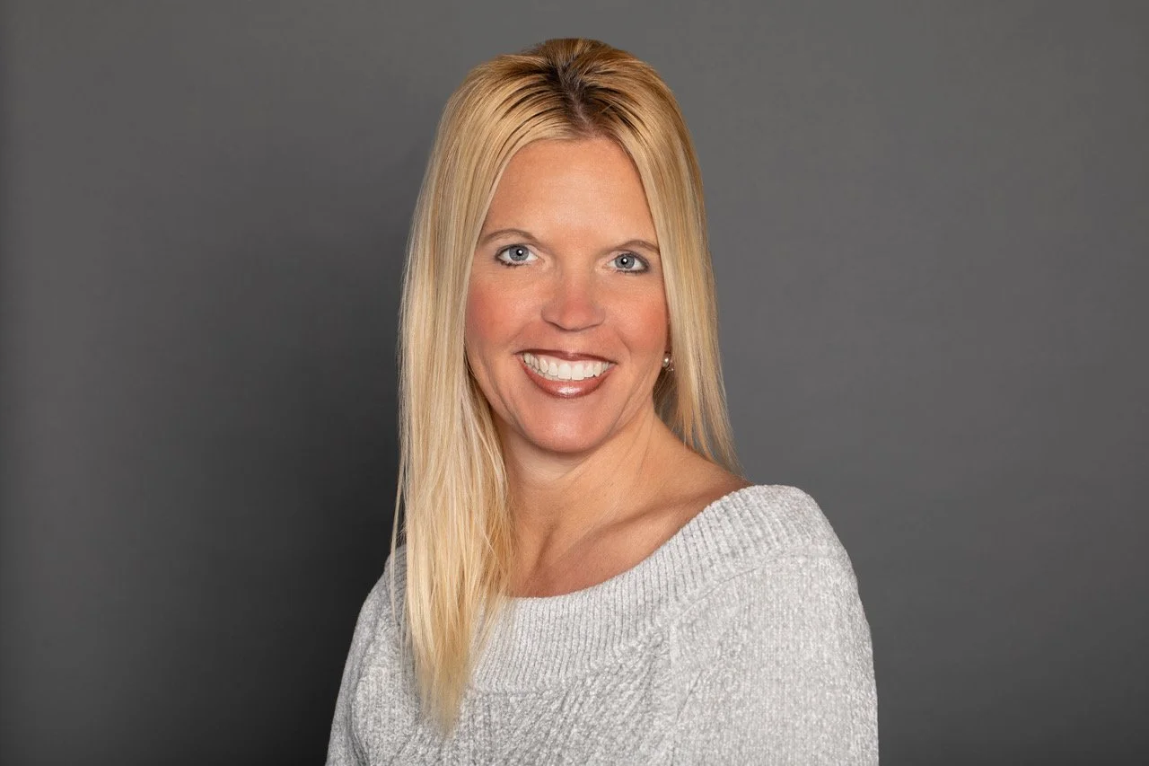 Headshot of a blonde woman with blue eyes, smiling, wearing a light gray sweater, against a dark gray background.