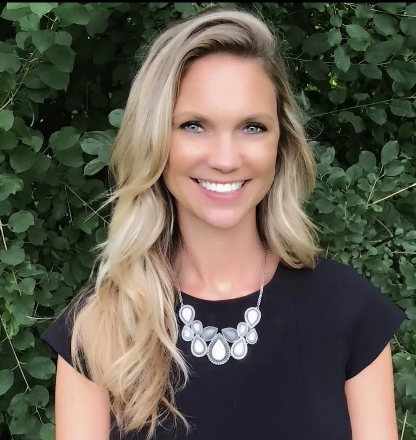 Young woman with blonde hair smiling, wearing a black top and a silver statement necklace, standing in front of green foliage.