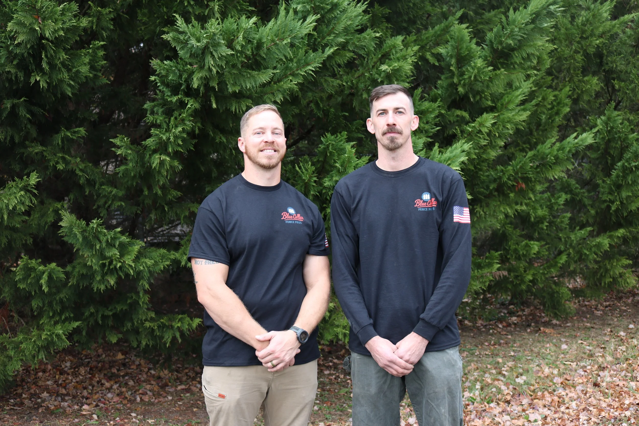 Two men standing outdoors in front of green pine trees, both wearing black long-sleeve shirts with patches and logos, one smiling and the other with a serious expression.