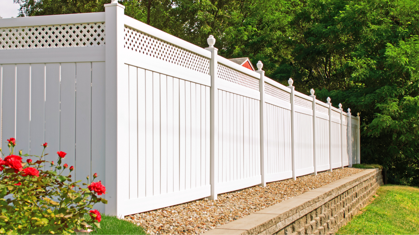 White vinyl privacy fence with decorative lattice top, set above a stone retaining wall, with green trees and red flowers in the background.