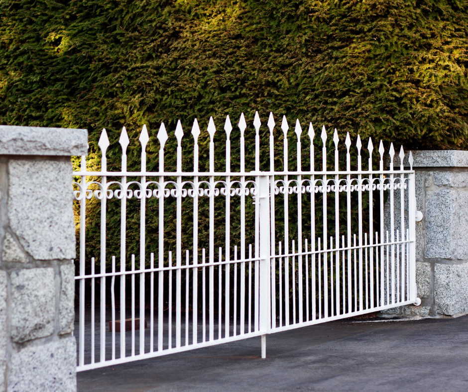 A white metal gate with pointed tips and decorative circles, flanked by stone pillars, with green bushes in the background.
