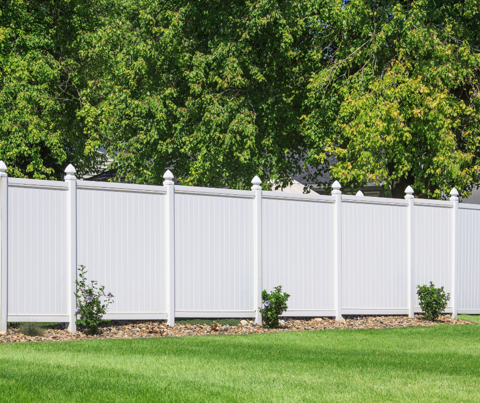 White vinyl privacy fence with small bushes and green grass in front, and leafy trees in the background.