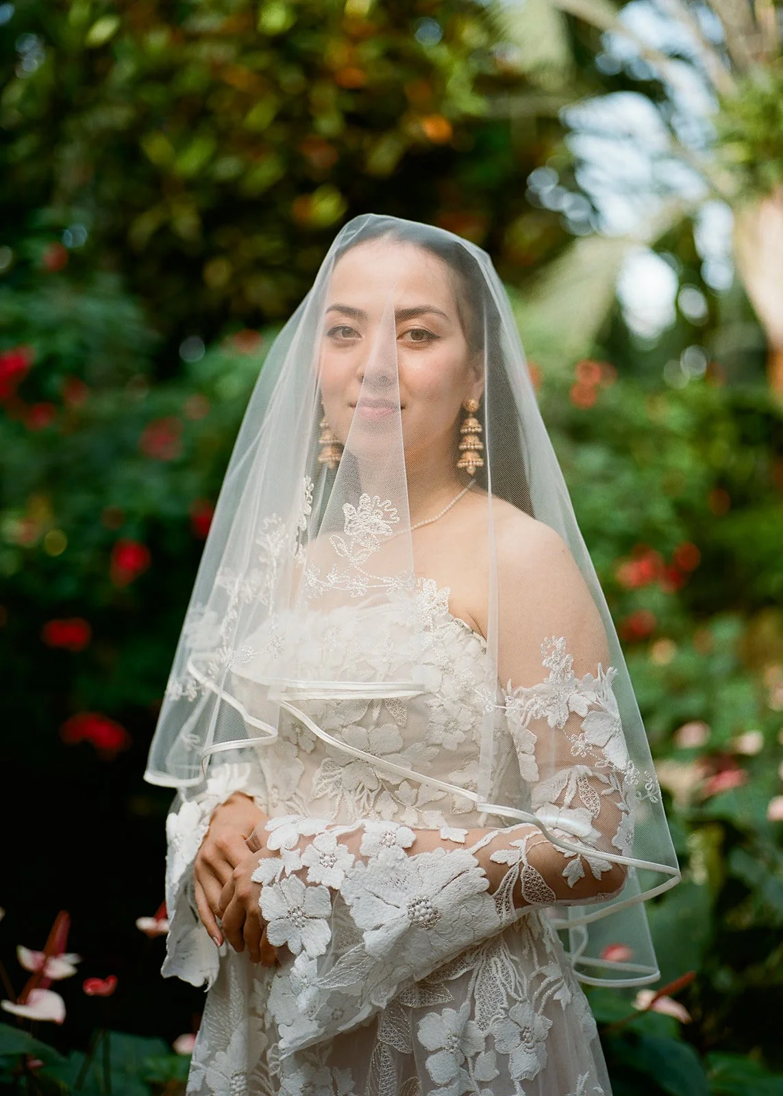 A woman in a wedding dress with lace details and floral embroidery, wearing a veil and jewelry, standing outdoors with greenery and red flowers in the background.