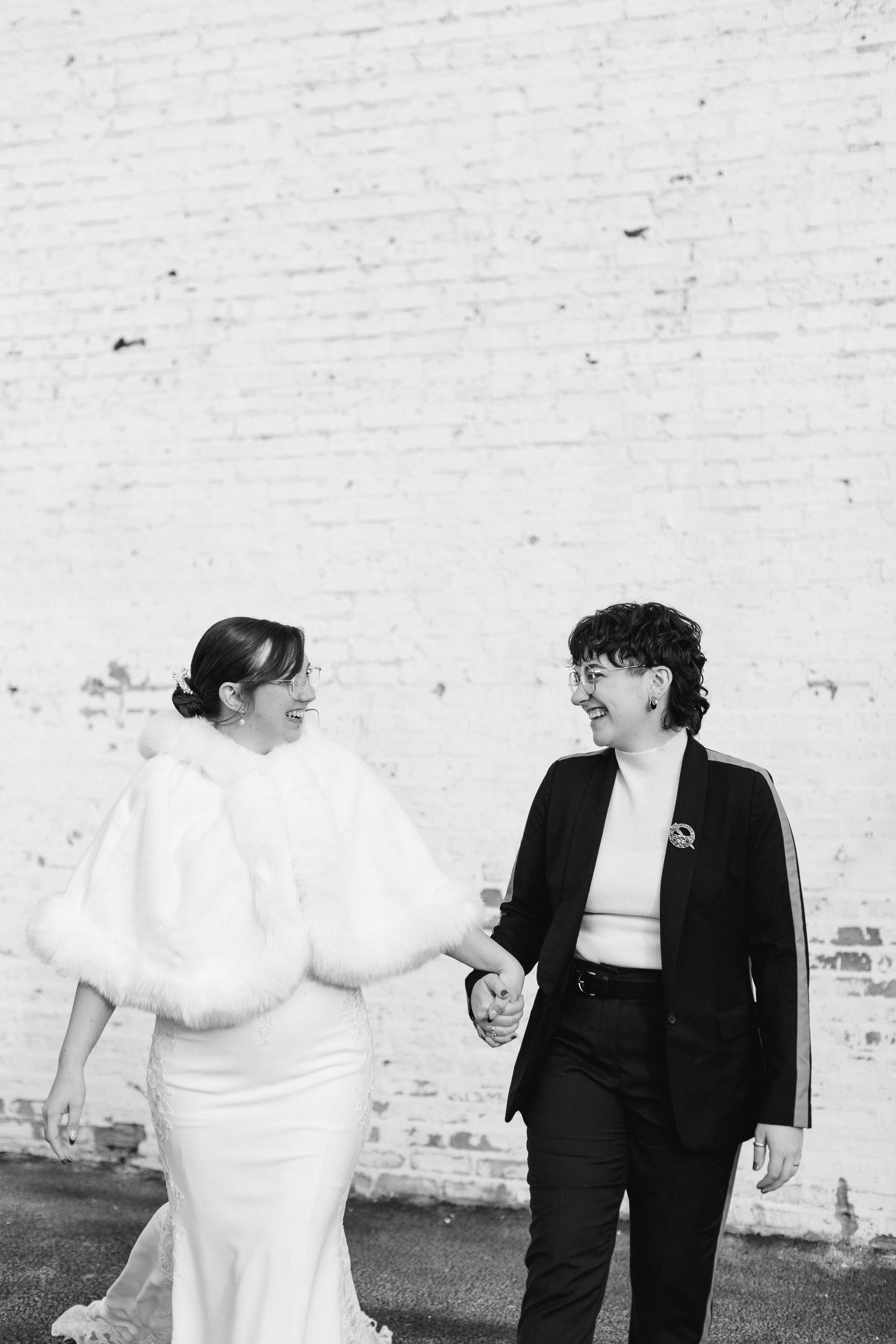 Black and white photo of two brides holding hands walking. Grace Coudal is a Chicago lesbian photographer specializing in portraits, product, food, lifestyle, events, + weddings. Analog film photography, queer-owned.