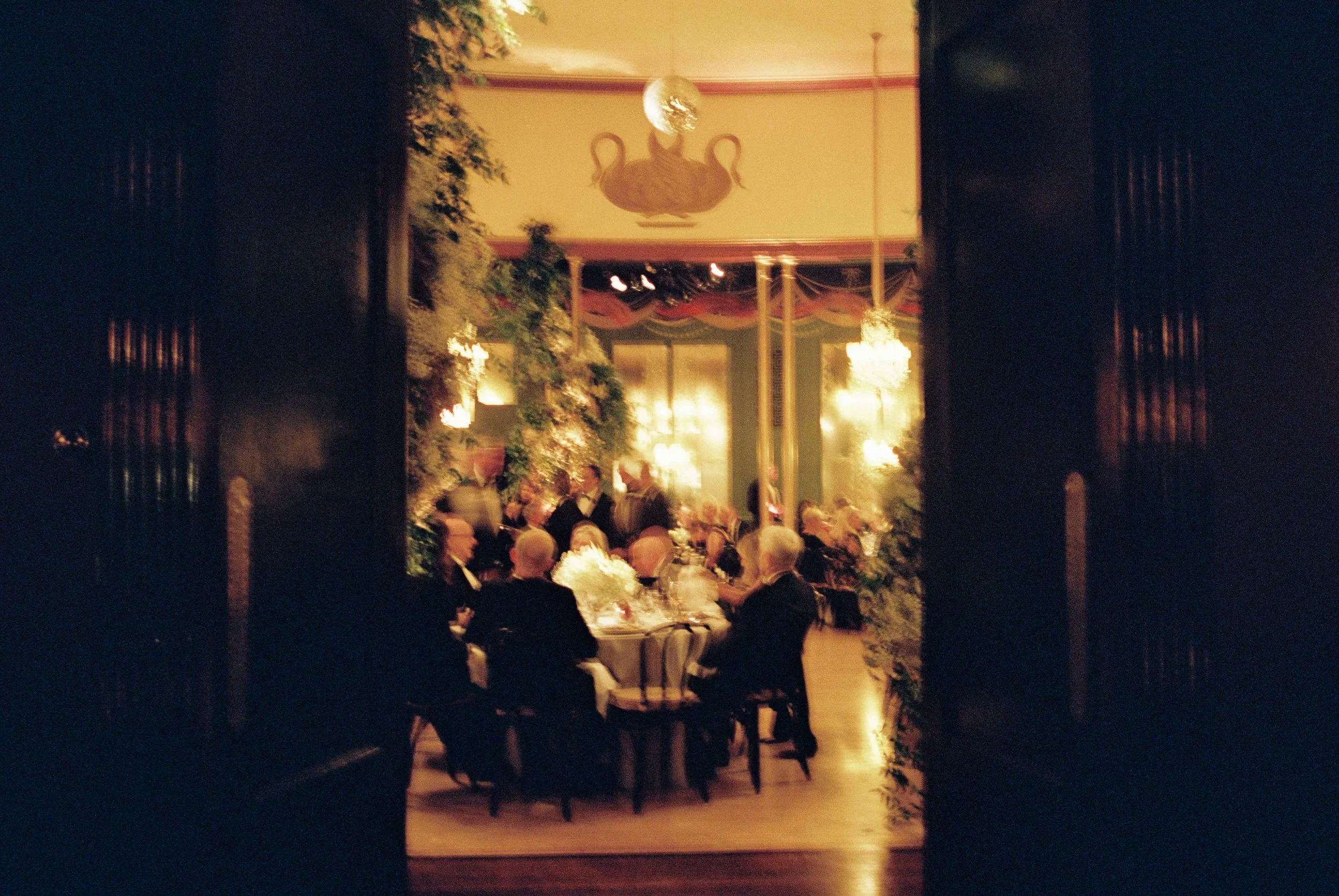 A view through a doorway into an elegant, dimly lit banquet room with decorated walls, chandeliers, and a gathering of formally dressed people seated around a table.
