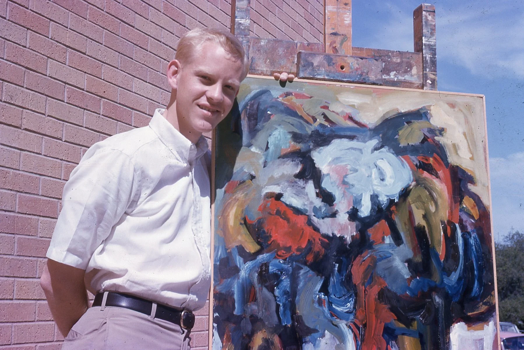 A young man with light-colored hair, wearing a white shirt, beige pants, and a black belt, standing outdoors next to an abstract portrait painting of a bulldog on a wooden easel against a brick wall, with a blue sky in the background.