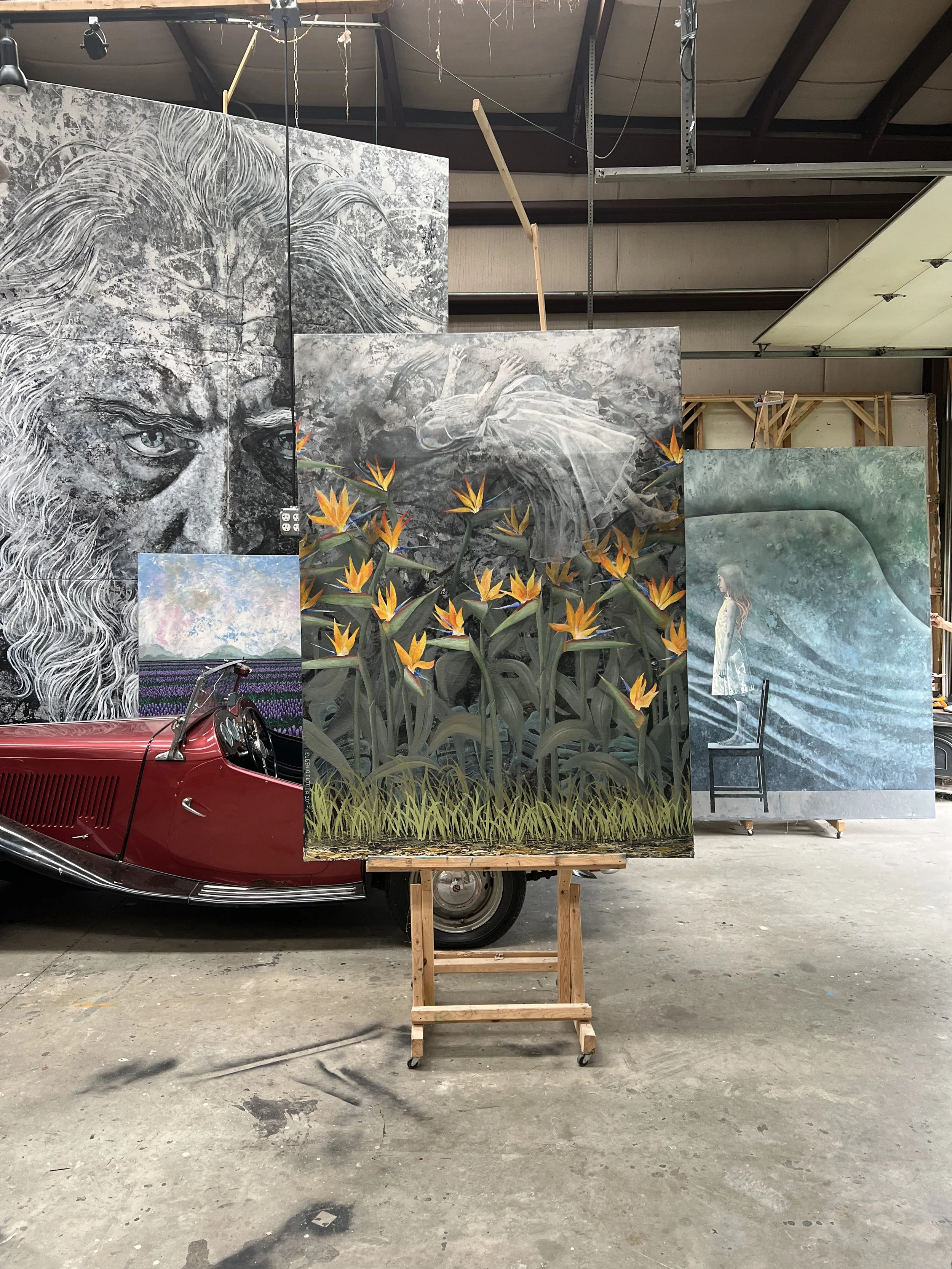 Diversas pinturas en un taller, incluyendo un retrato en blanco y negro con ojos azules, una escena de una mujer en un río, un campo de flores y una obra abstracta con una mujer de perfil, junto a un coche clásico rojo en el fondo.