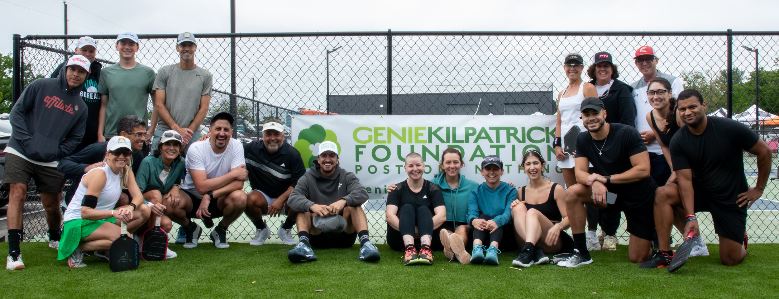 Group of people posing behind a banner at a tennis court, with a fence and cloudy sky in the background, participating in a Genie Kilpatrick Foundation charity event.