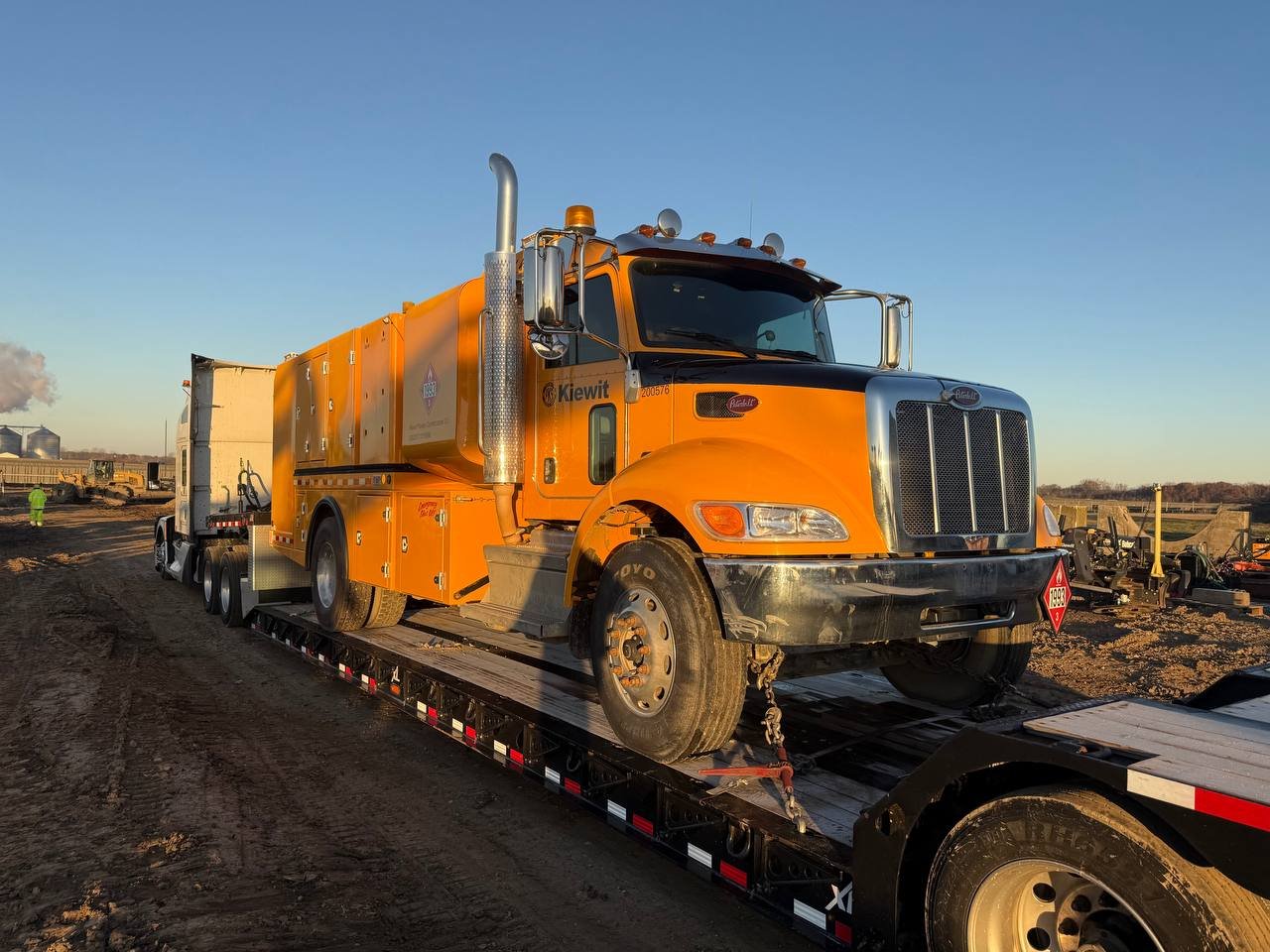Orange utility truck loaded on a flatbed trailer in an outdoor construction or industrial site during daytime.
