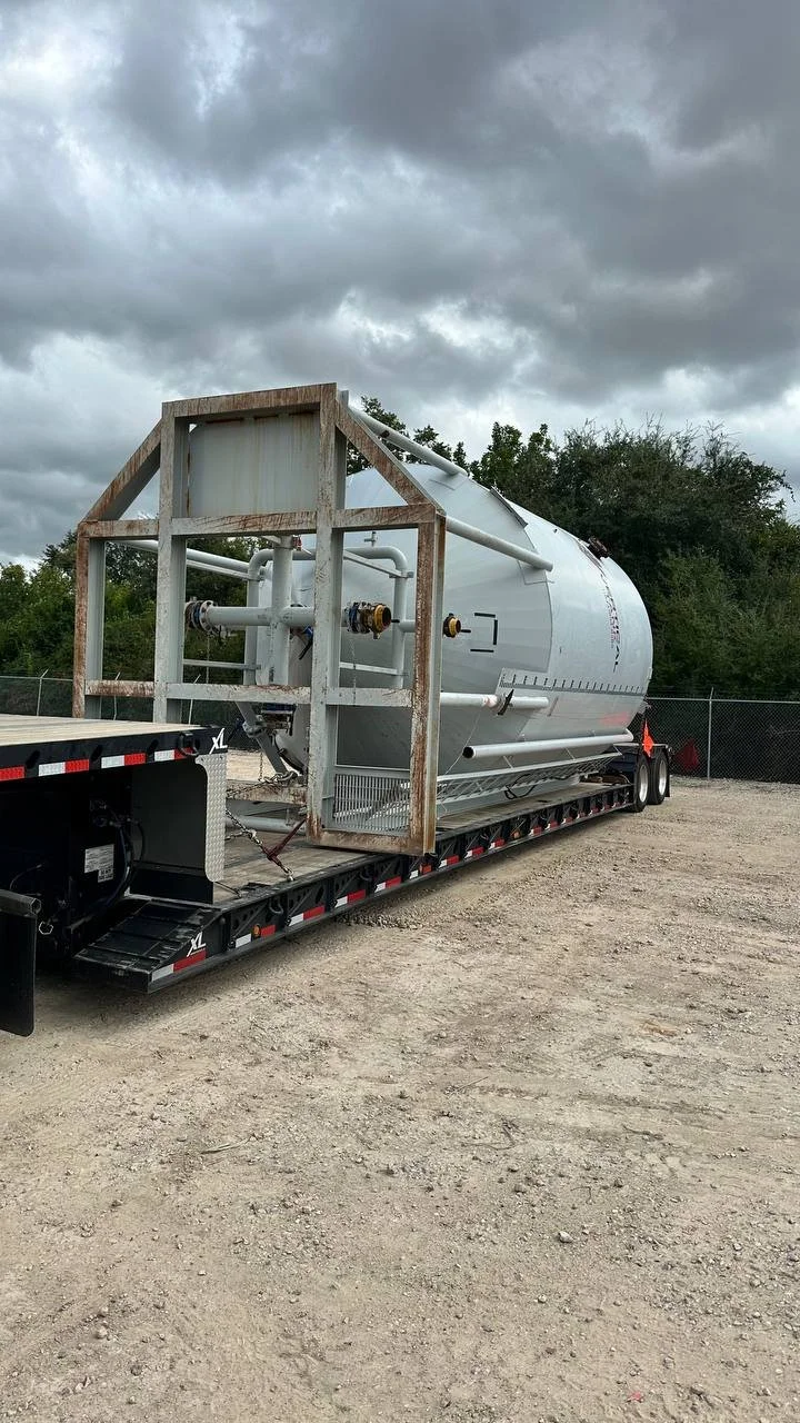A large cylindrical tank, possibly for liquids or gases, mounted on a flatbed trailer at an outdoor location with a dirt ground and a cloudy sky.
