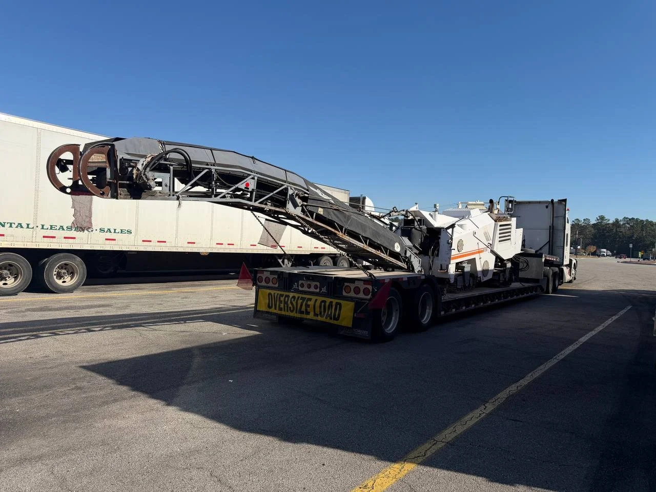 Large construction or industrial machine with an 'Oversize Load' banner on the back, parked in a lot with a truck in the background, under a clear blue sky.