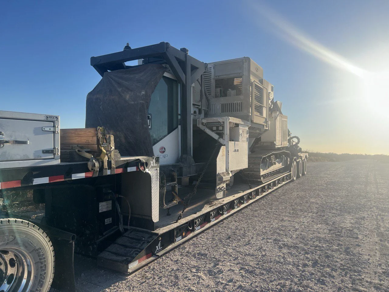 A tracked piece of construction or mining equipment loaded onto a flatbed trailer, parked on a dirt surface at sunset.
