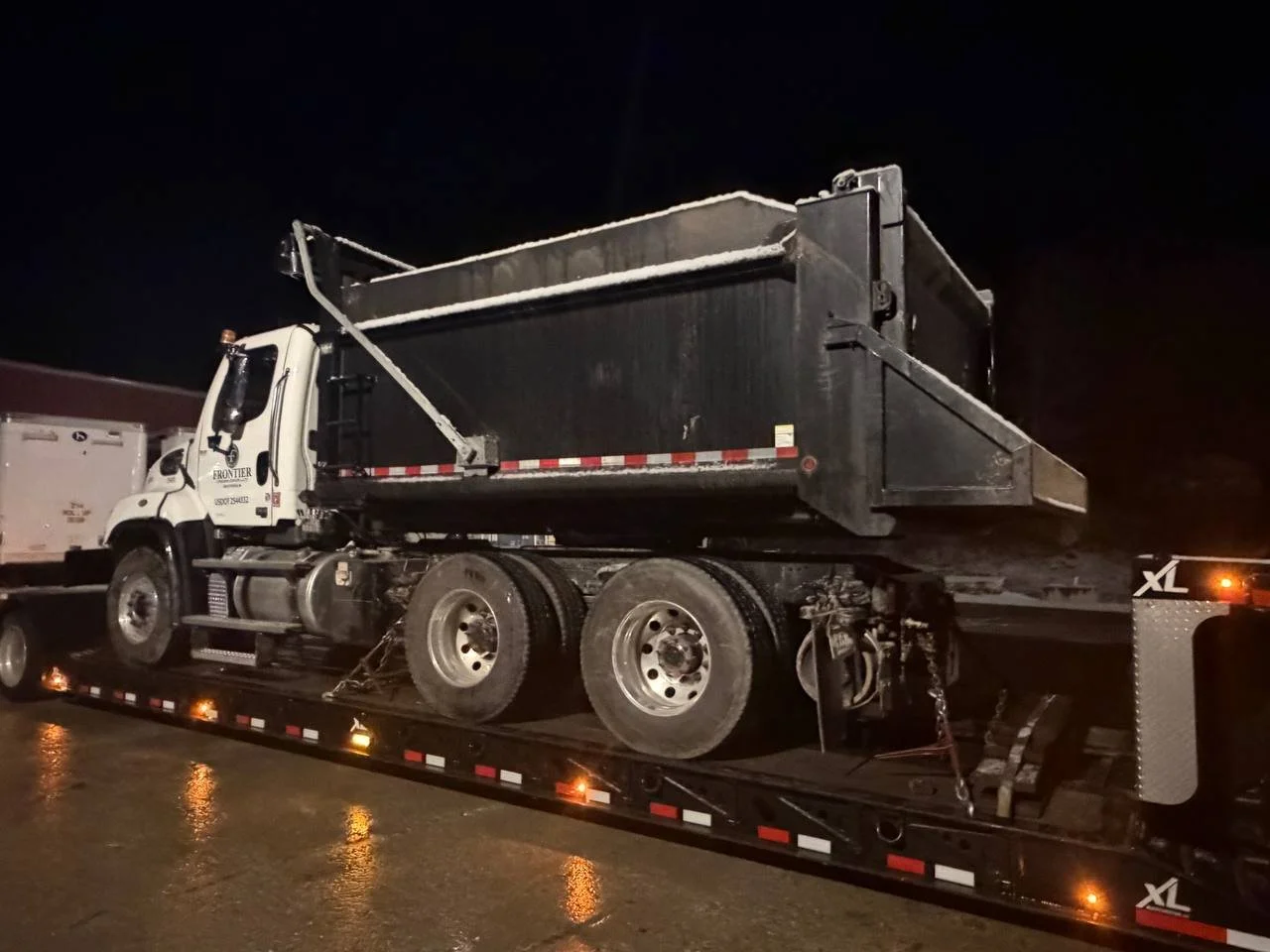 A dump truck loaded on a flatbed trailer at night, with snow on top of the truck bed, in a wet environment.