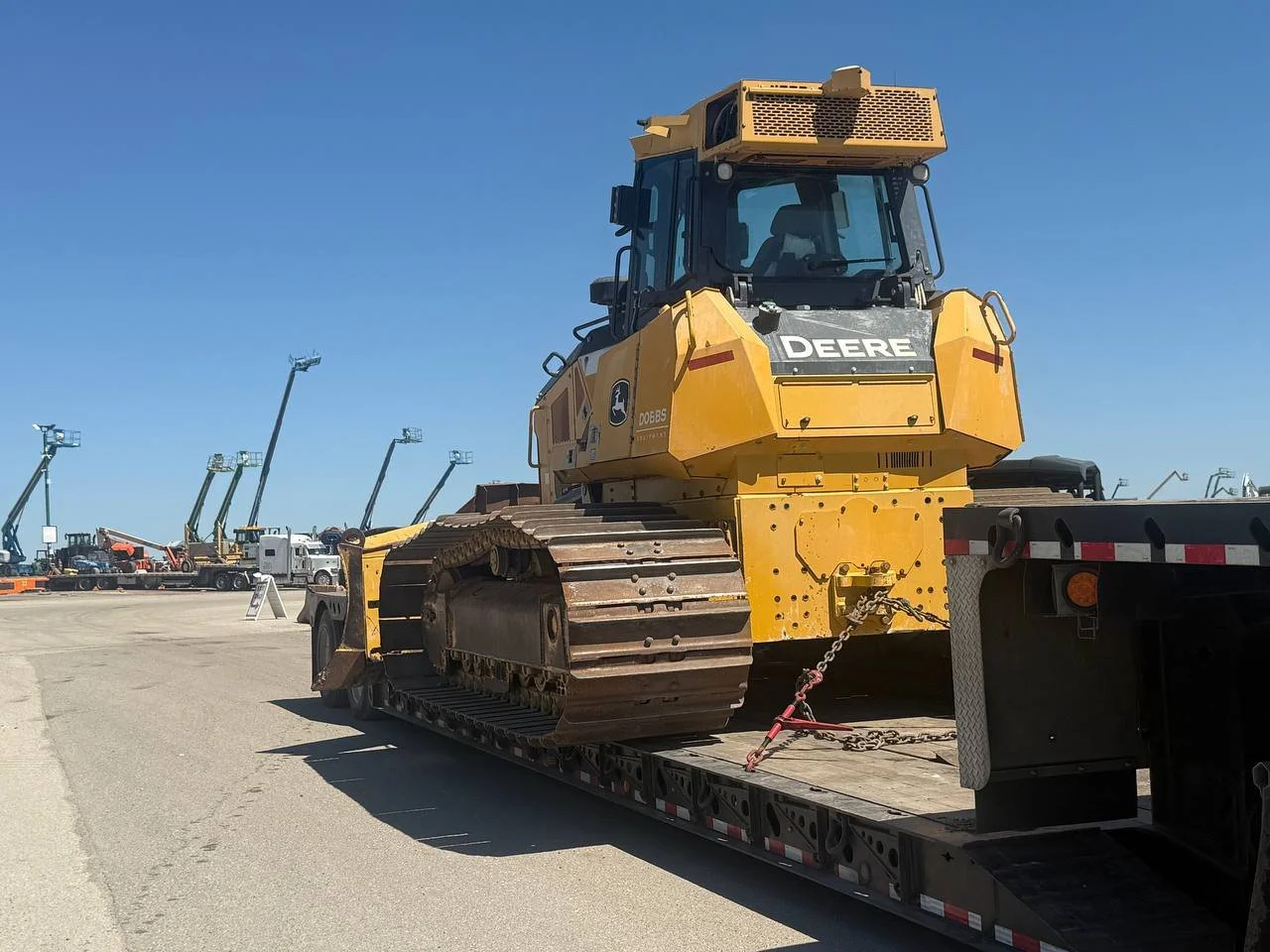 Large yellow John Deere bulldozer on flatbed trailer in an outdoor lot with construction equipment in the background.